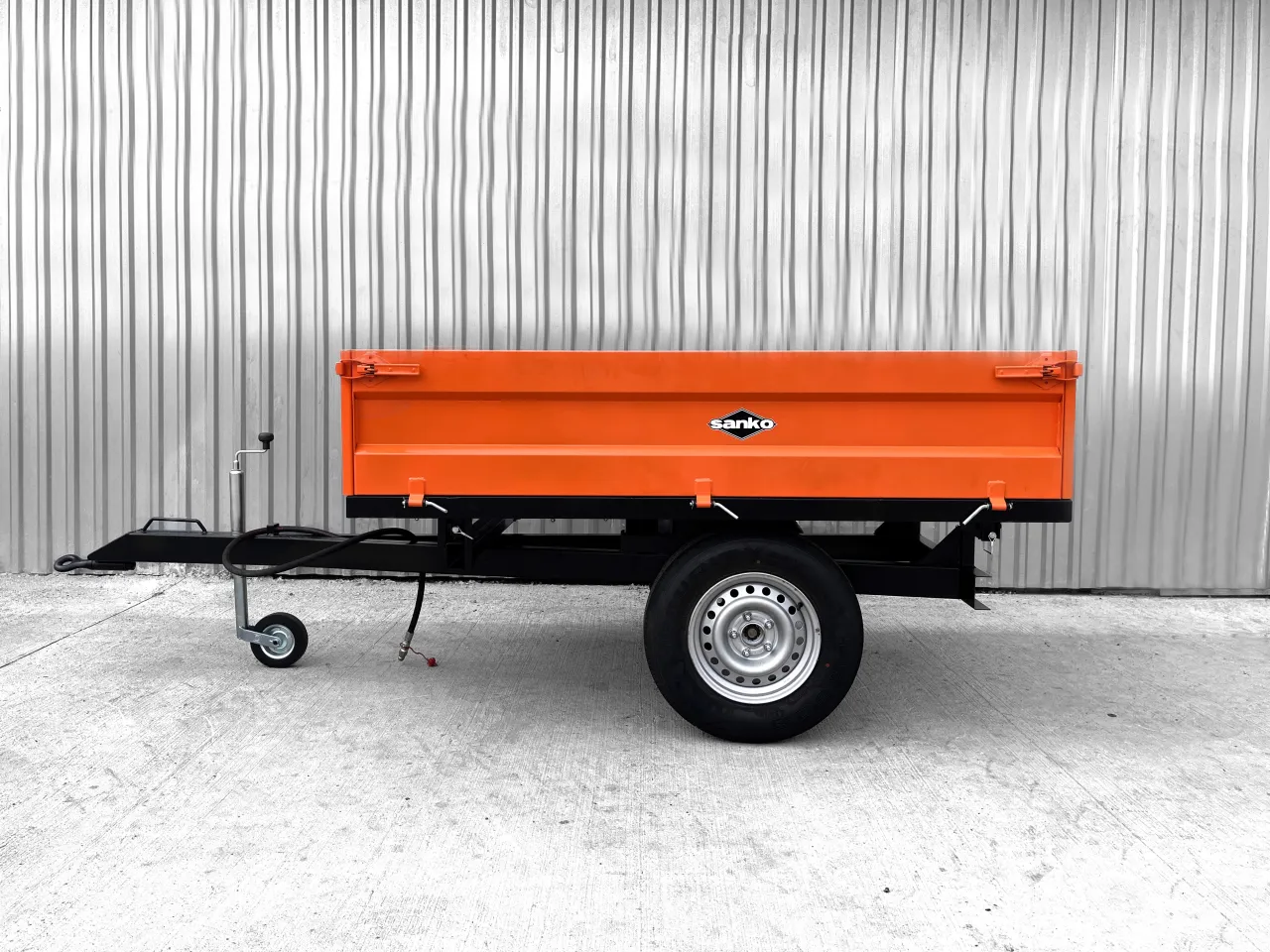 Pomarańczowa przyczepka do traktorka z czarną ramą i kołem, gotowa do pracy w ogrodzie.