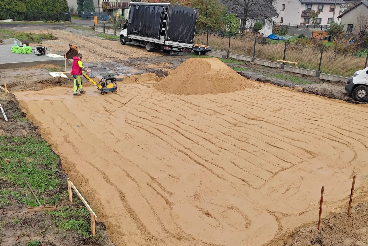 Pracownik zagęszcza piasek zagęszczarką. To kluczowy etap wymiany gruntu pod płytę fundamentową, zapewniający stabilność.