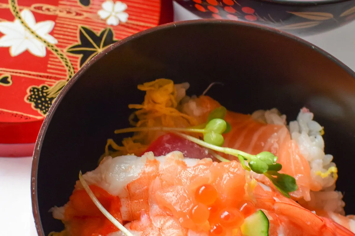 Chirashi zushi z krewetkami, łososiem i ikrą łososia, podane w czarnej misce, obok ozdobnego czerwonego pudełka.