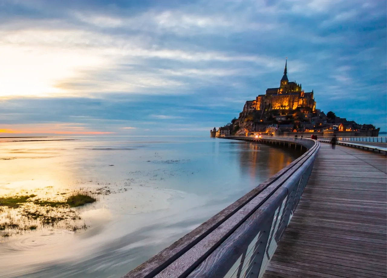 Mont Saint-Michel, najpopularniejsze sanktuarium we Francji, majestatycznie wznosi się nad wodą o zachodzie słońca. Drewniana kładka prowadzi do tego cudu architektury.