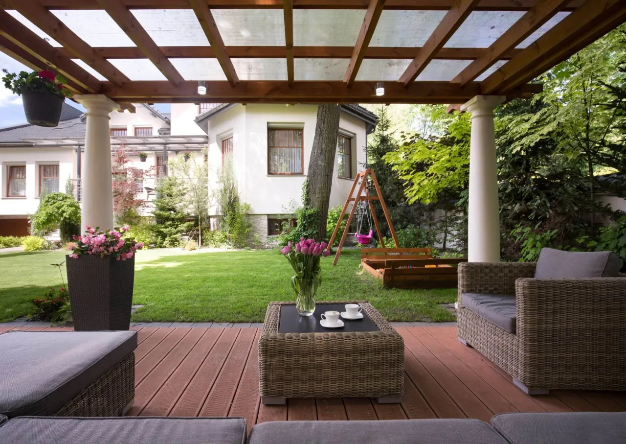 Drewniana altana z przezroczystym dachem, rattanowe meble i bujna zieleń tworzą idealne miejsce na relaks. Co na dach altany? Przezroczyste płyty chronią przed słońcem i deszczem.