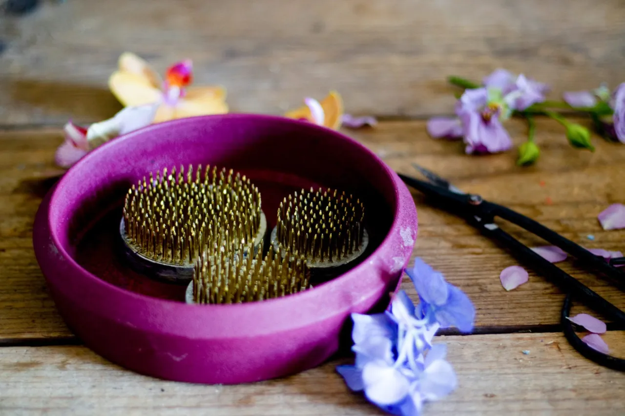 Przygotowania do układania kwiatów w gąbce florystycznej. Na drewnianym blacie leżą kenzany w fioletowej misie, obok nożyczki i luźne płatki kwiatów.