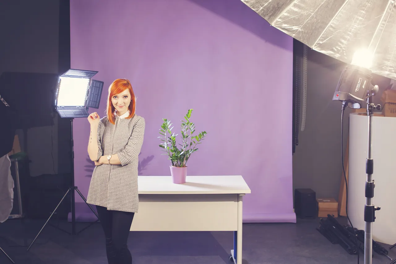 Kobieta z rudymi włosami pozuje do zdjęcia produktowego na fioletowym tle. Widać lampy studyjne i roślinę w doniczce.