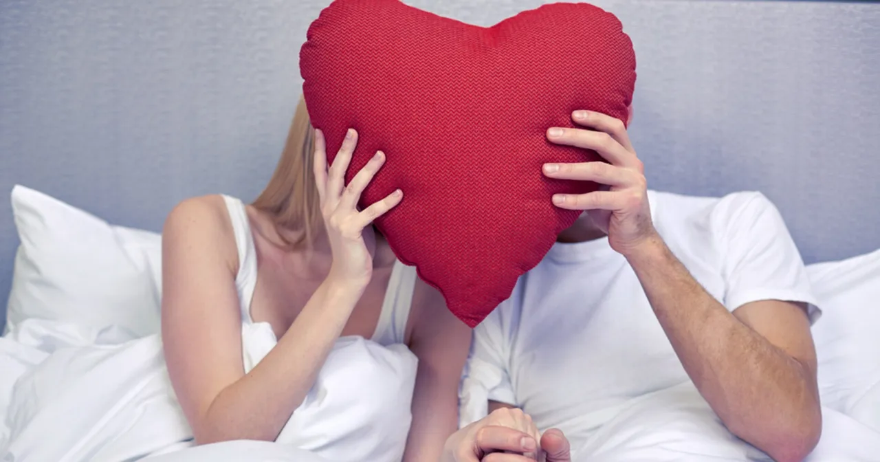 Para, trzymając się za ręce, ukrywa twarze za czerwoną poduszką w kształcie serca. Szeptem 