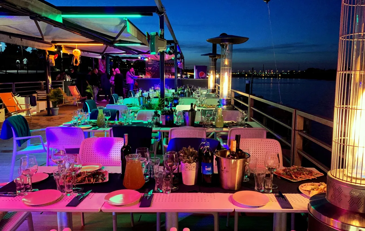 Barka warszawska dzień i noc, oświetlona neonami, z nakrytymi stołami i gośćmi przy barze.