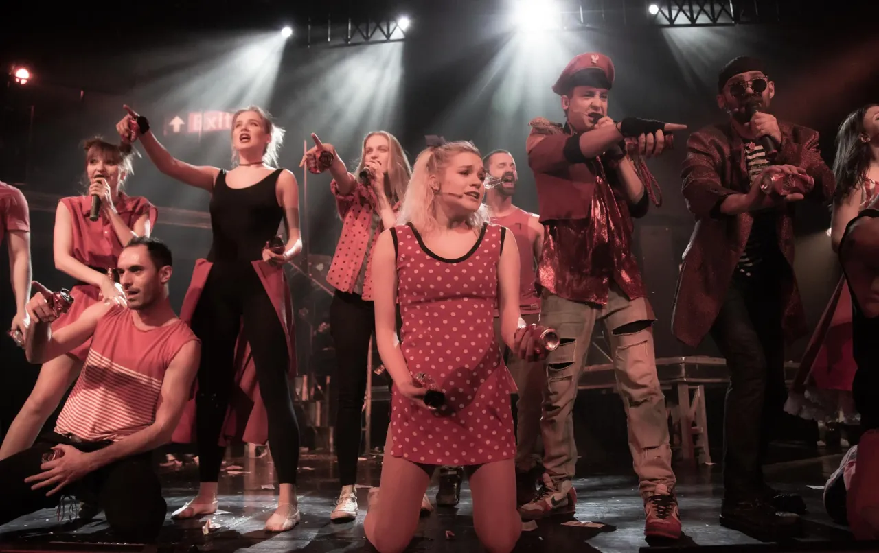 Aktorzy na scenie, w czerwonych strojach, śpiewają i tańczą. Czy to część musicalu 