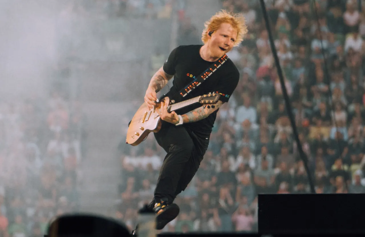 Ed Sheeran skacze na scenie, grając na gitarze. Fani zastanawiają się, ile Ed Sheeran bierze za koncert, widząc jego energię.