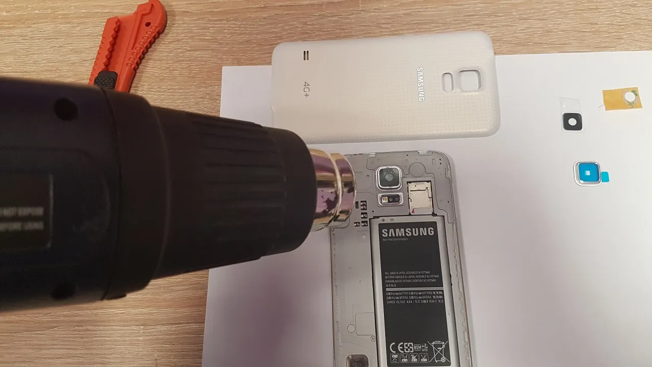 Ciepłe powietrze z opalarki ułatwia demontaż, gdy chcesz wymienić szybkę aparatu w telefonie. Obok leży rozkręcony Samsung.