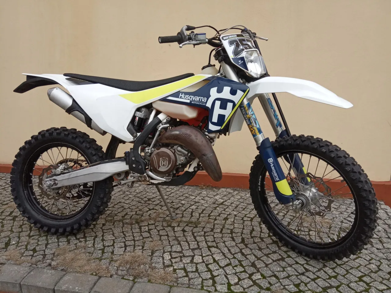 Husqvarna 125 2t gotowy do akcji na brukowanej nawierzchni. Biało-niebieski motocykl z żółtymi akcentami i numerem 25.