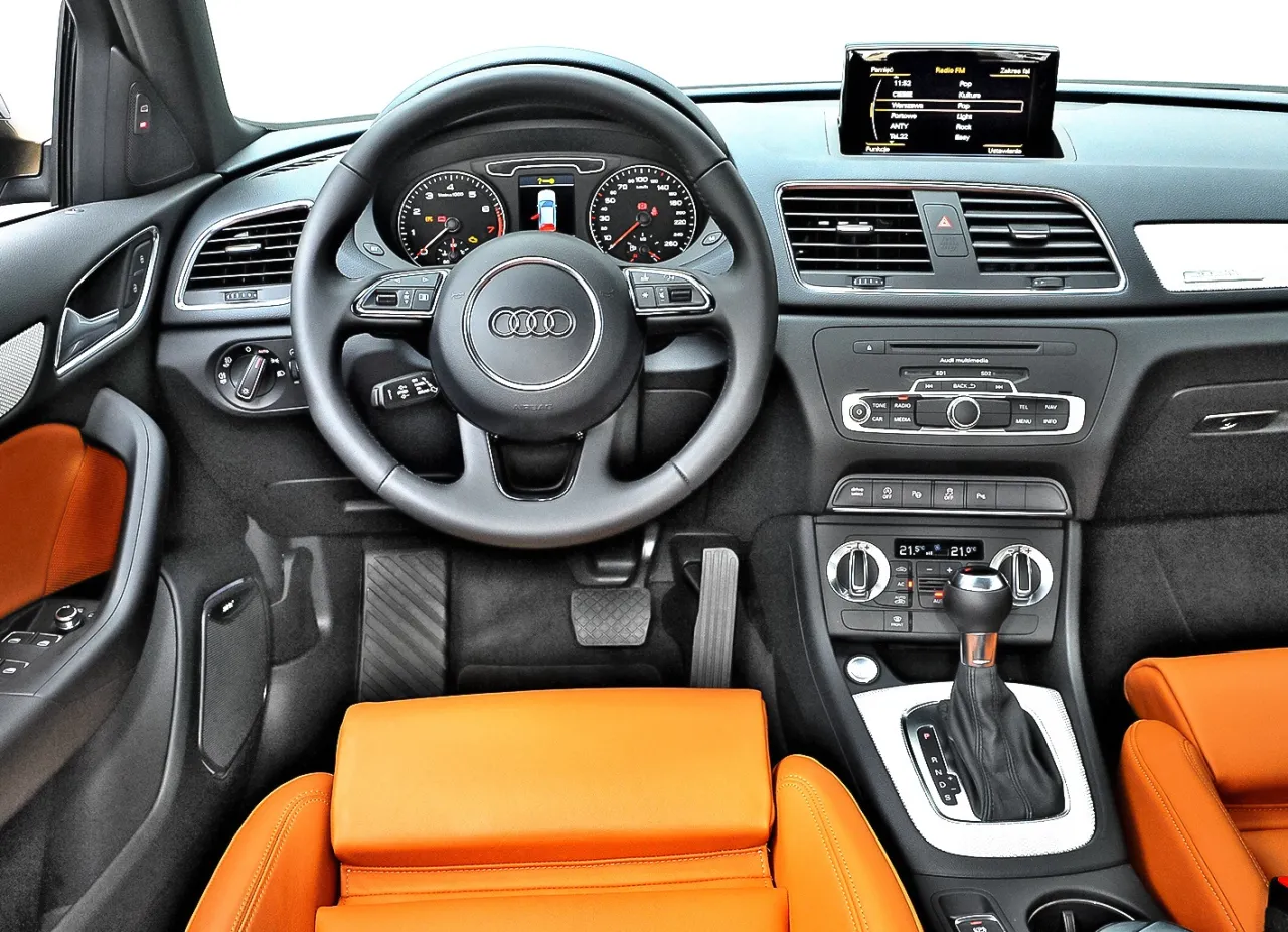 Wnętrze Audi Q3 z pomarańczową tapicerką. Zastanawiasz się, które audi wybrać? Ten model ma sportową kierownicę i nowoczesny panel.
