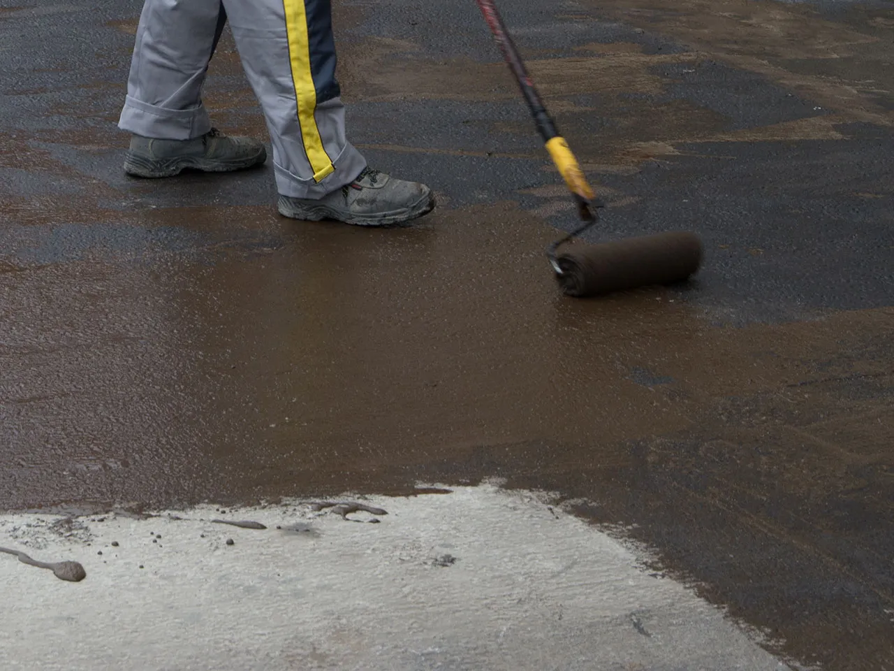 Pracownik wałkiem rozprowadza ciemną masę na betonowej wylewce. Jaki beton na wylewkę? To pytanie zadaje sobie wielu budowlańc&oacute;w.
