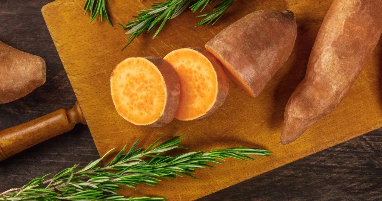 Na desce do krojenia leżą pomarańczowe bataty, pokrojone w plastry, obok gałązki rozmarynu. Czy bataty są zdrowe? Tak, to pyszne i zdrowe warzywo!