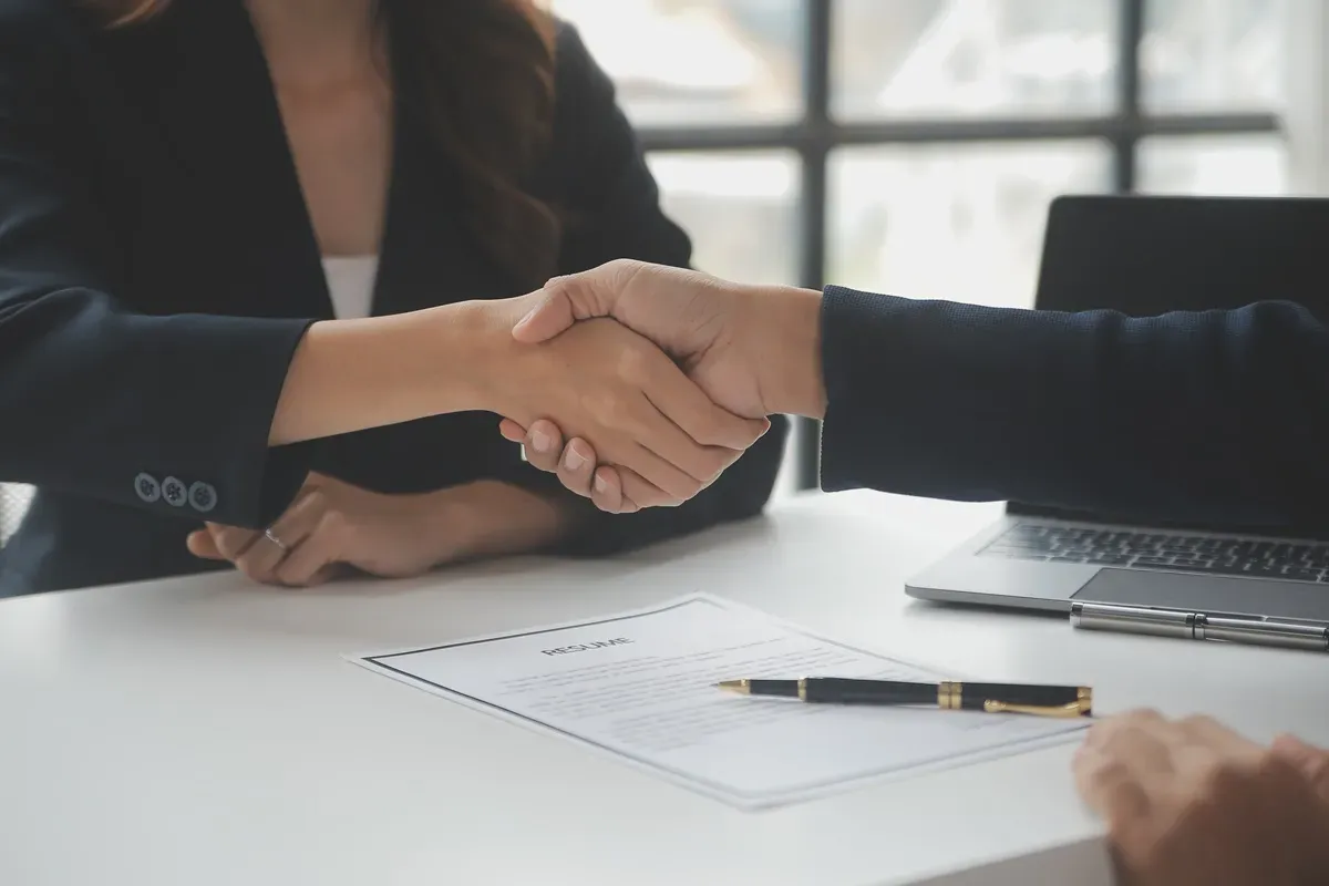 Uścisk dłoni po rozmowie kwalifikacyjnej. Czy po stażu jest obowiązek zatrudnienia? Na stole CV i długopis.