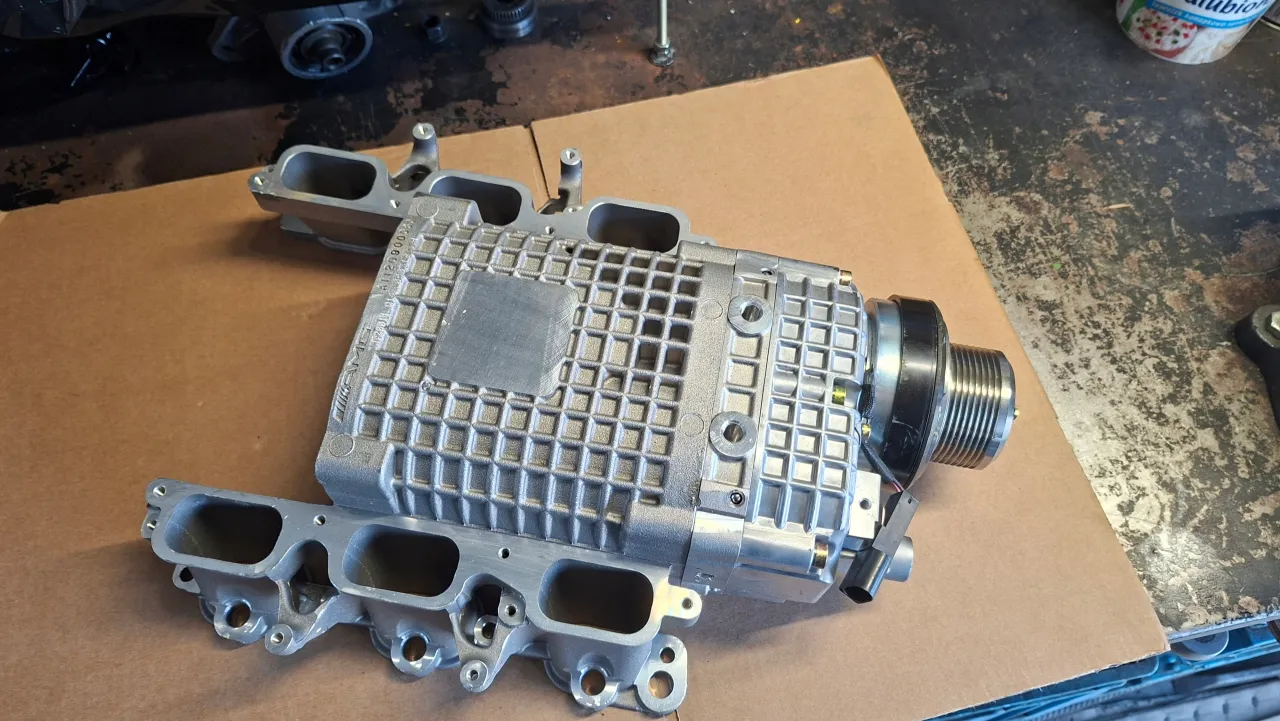 Srebrna sprężarka mechaniczna z kolektorem dolotowym, część do Mercedes Benz C32 AMG, leży na brązowym kartonie.