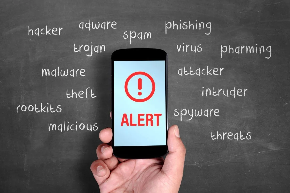Dłoń trzyma telefon z napisem ALERT. Wok&oacute;ł na tablicy wypisano zagrożenia: hacker, adware, trojan, malware, rootkits, malicious, spam, phishing, virus, attacker, intruder, spyware, pharming, threats.
