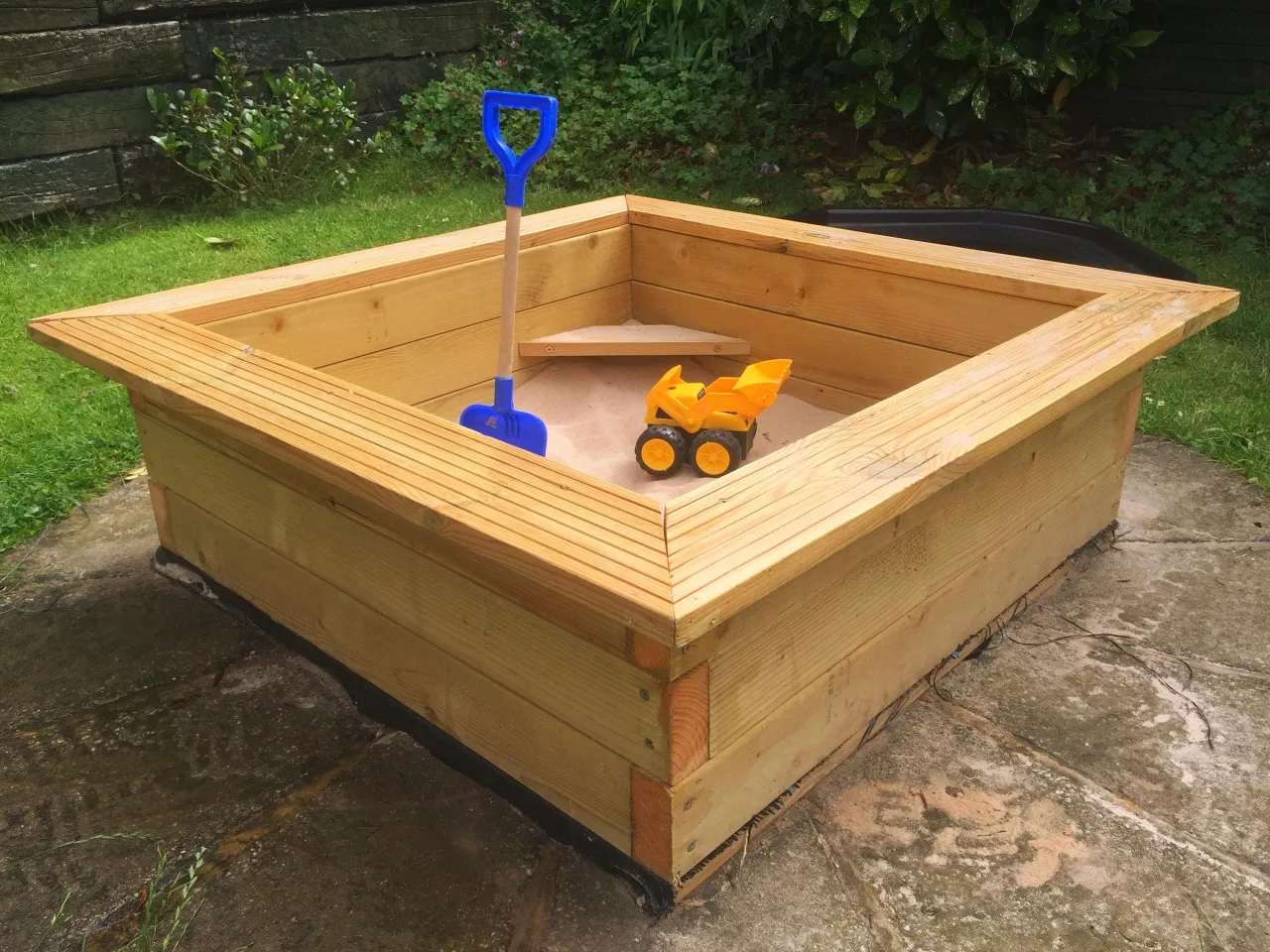 Gotowa piaskownica z drewna, z niebieską łopatką i żółtą koparką. Idealne miejsce do zabawy, pokazujące, jak zrobić piaskownicę.