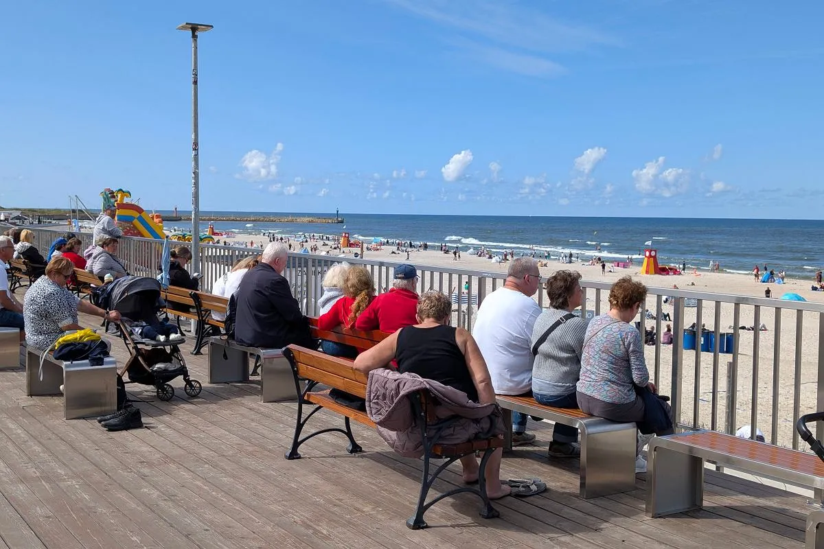 Ludzie siedzą na ławkach, patrząc na plażę i morze. Czy zastanawiasz się, ile mieszkańców ma Łeba?