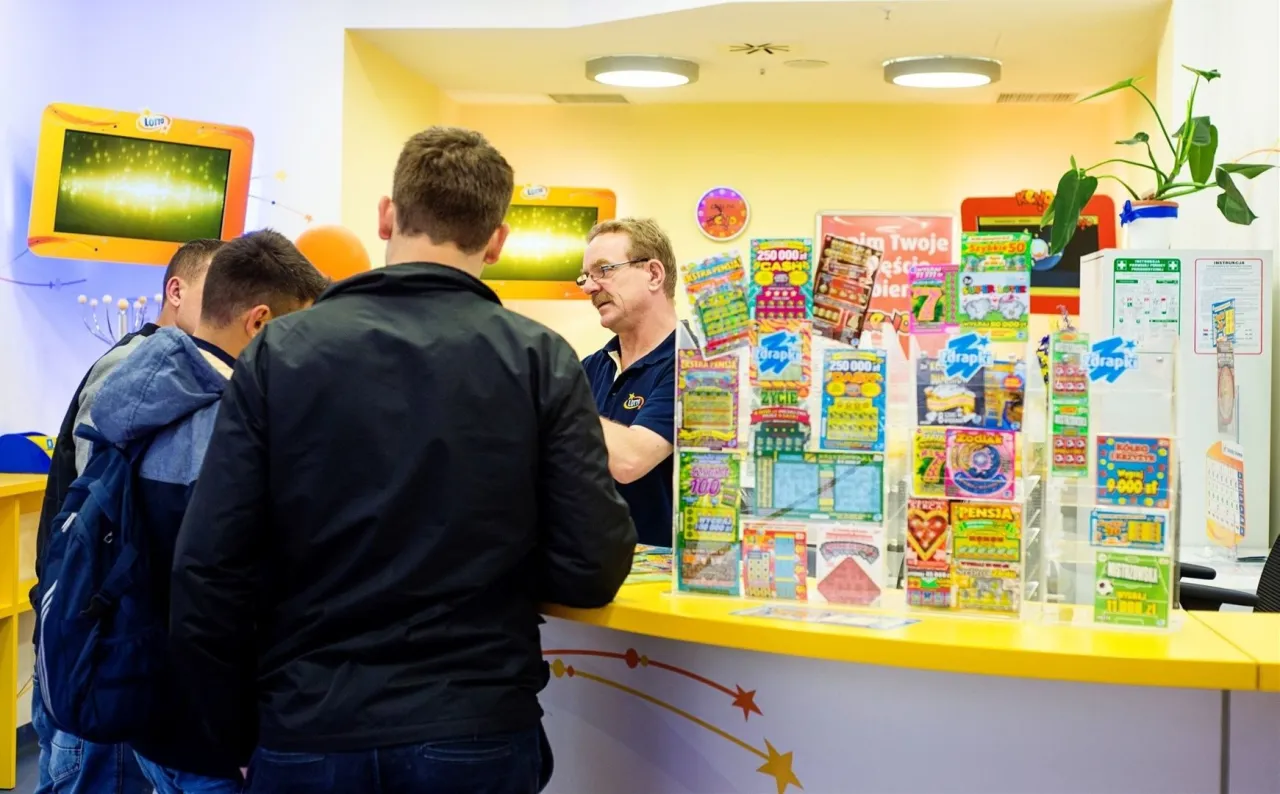 Mężczyźni stoją przy ladzie z licznymi zdrapkami. Czy wygraną zdrapki można wymienić wszedzie? Sprzedawca w okularach odpowiada na pytania.