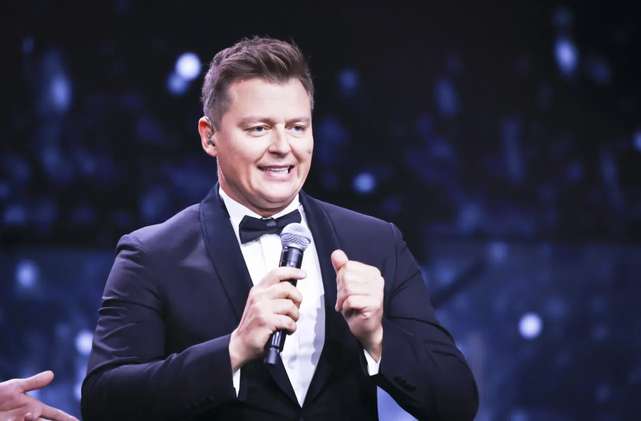 Rafał Brzozowski w garniturze z mikrofonem, uśmiechnięty. Czy to odpowiedź na pytanie dlaczego Rafał Brzozowski odszedł z Koła Fortuny?