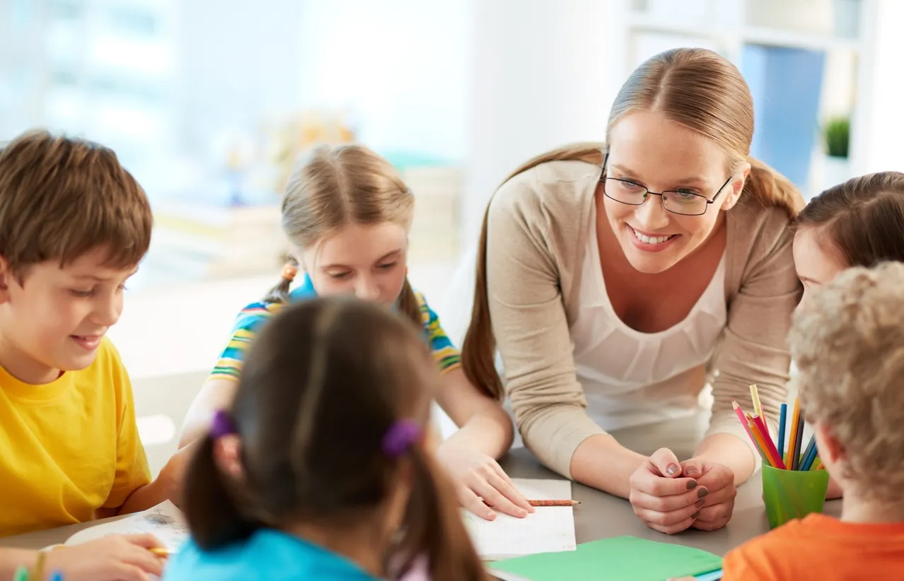 Nauczycielka z uśmiechem pomaga dzieciom w nauce. Dowiedz się, jak zdobyć uprawnienia pedagogiczne, by móc wspierać rozwój najmłodszych.