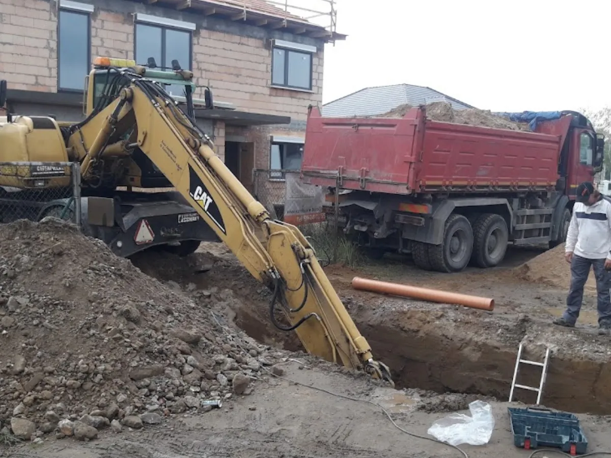 Żółta minikoparka CAT kopie rów. Obok stoi czerwona wywrotka z piaskiem. Trudno określić, ile pali minikoparka na godzinę, ale praca wre.