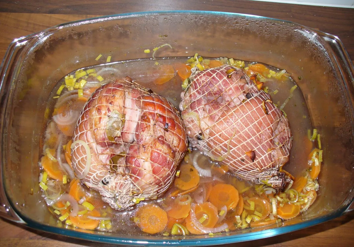 Dwie golonki w siatce, gotowe do peklowania, w naczyniu żaroodpornym z marchewką i porem.