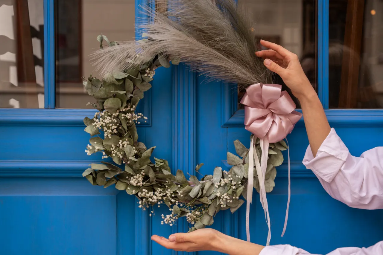 Jak zrobić wianek dekoracyjny? Delikatny wianek z liści i drobnych białych kwiat&oacute;w, ozdobiony pudrowym, satynowym  kokardą, wisi na niebieskich drzwiach.