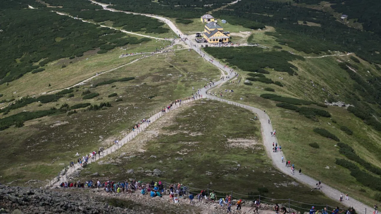 Tłum turyst&oacute;w na kamienistym szlaku, kt&oacute;ry szlak na Śnieżkę jest najtrudniejszy? Widok z g&oacute;ry na g&oacute;rzysty teren z zielenią i budynkami.