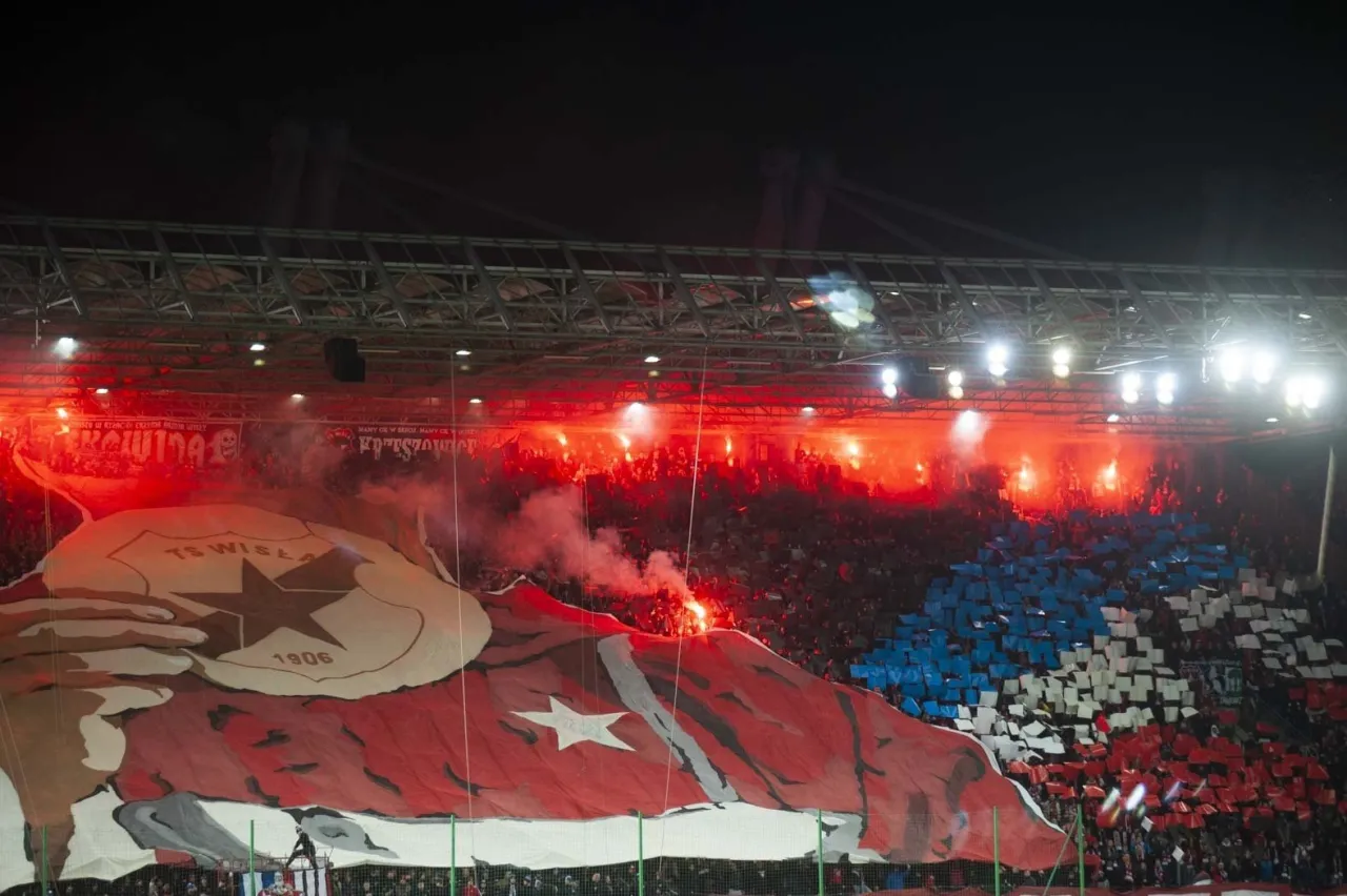 Oprawa meczu derbowego: kibice z flagami i racami tworzą czerwono-biało-niebieską mozaikę. To pokazuje, co to są derby w piłce nożnej.