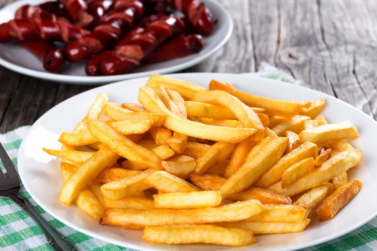 Złociste frytki na talerzu, idealne po tym, jak dowiesz się, jak przygotować ziemniaki na frytki. Obok talerz z kiełbaskami.