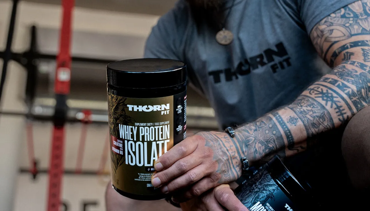 Mężczyzna z tatuażami trzyma opakowanie odżywki białkowej Thorn Fit. Idealne białko po treningu dla regeneracji mięśni.