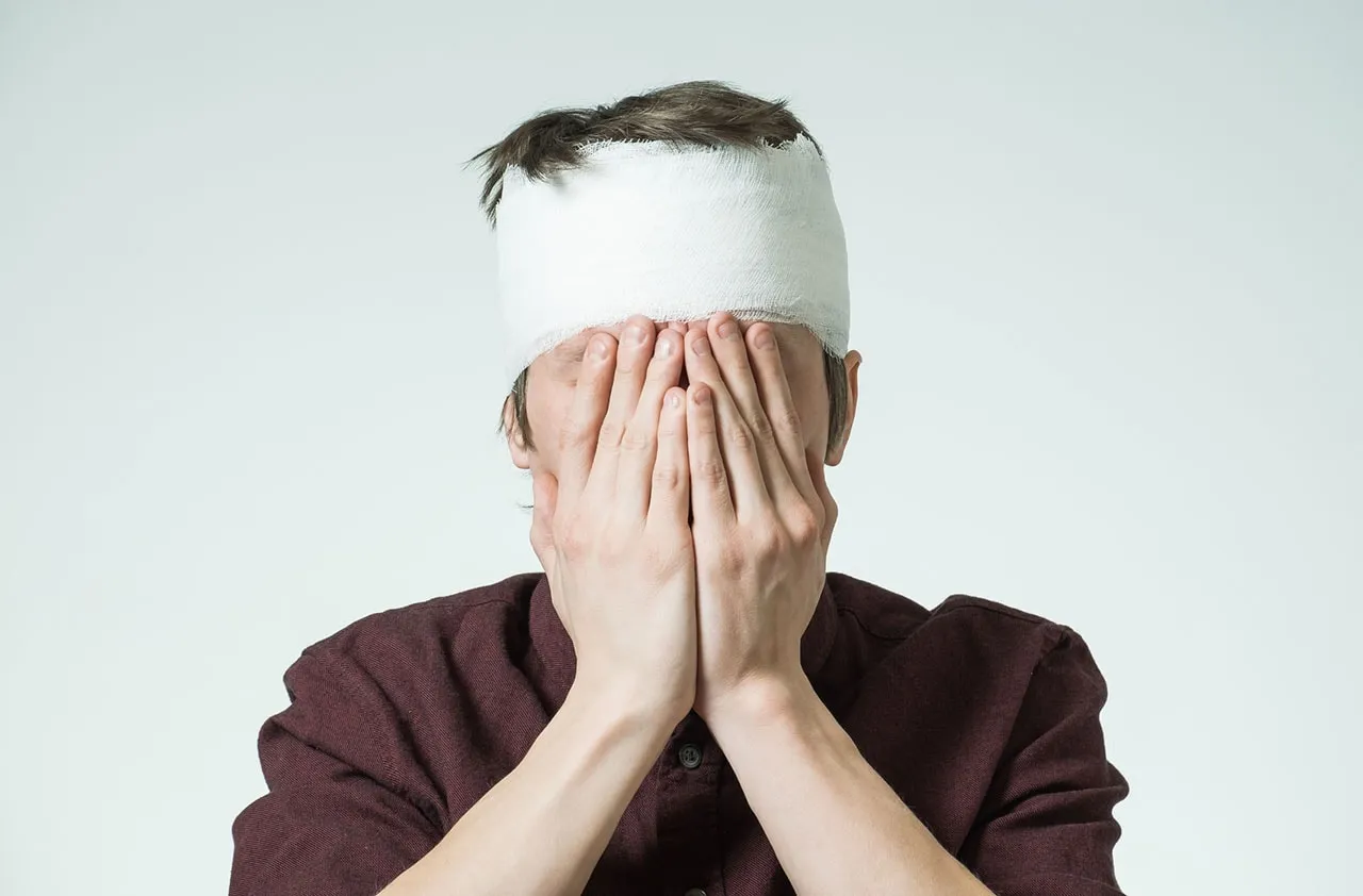 Mężczyzna z zabandażowaną głową, zakrywający twarz dłońmi, jakby po mocnym uderzeniu w głowę.