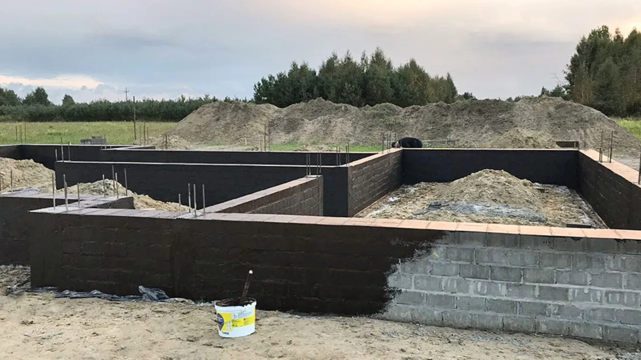 Fundamenty domu w budowie, ciemną masą izolacyjną. Widać wiadro z materiałem, czym smarować fundamenty.
