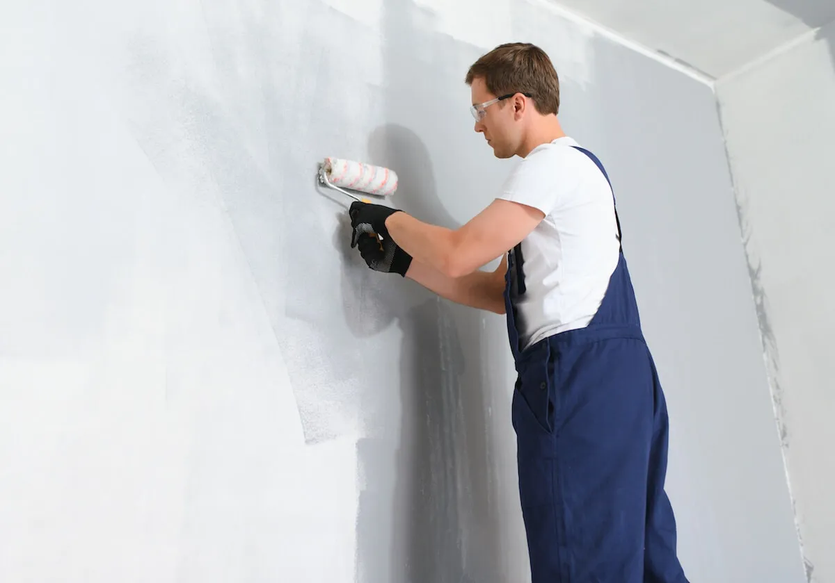 Mężczyzna w ogrodniczkach maluje ścianę wałkiem. Czy gładź można kłaść na farbę? To pytanie zadaje sobie wielu remontujących.