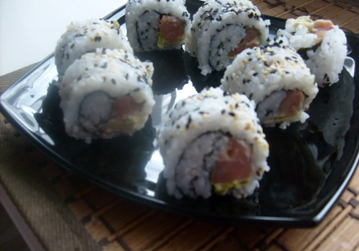 Uramaki co to? Rolki sushi z ryżem na zewnątrz, posypane sezamem, z kawałkami ryby i awokado w środku, podane na czarnym talerzu.