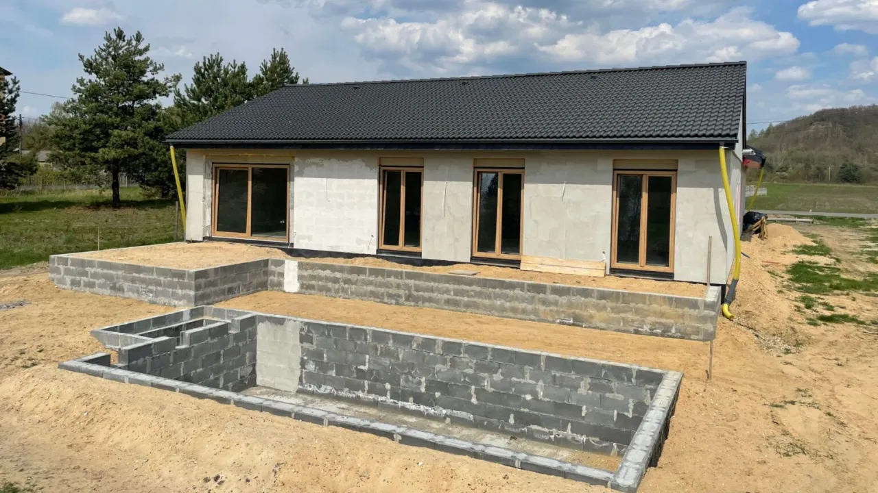 Budowa domu w toku: wzniesiony budynek z dachem i oknami, fundamenty z bloczków betonowych, piasek wokół.