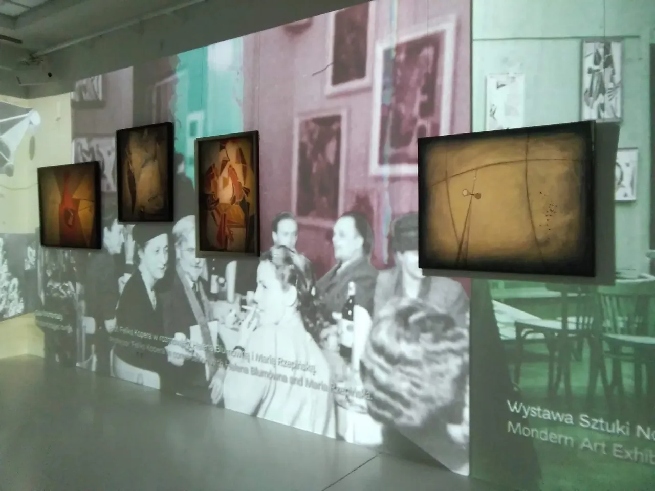 Wystawa sztuki nowoczesnej w jednym z muzeów sztuki w Polsce. Obrazy wiszą na tle projekcji historycznych zdjęć.