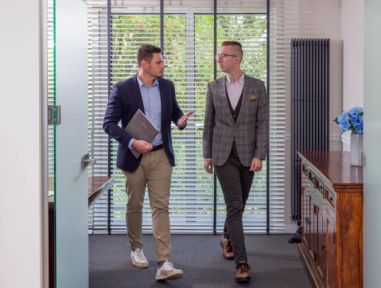 Dwóch mężczyzn w garniturach rozmawia, jeden trzyma teczkę. Dyskutują, jak zostać prawnikiem bez aplikacji, w nowoczesnym biurze.