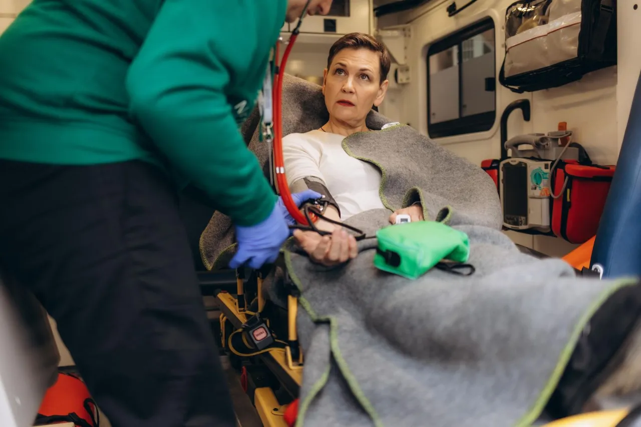 Kobieta w karetce, owinięta kocem, czeka na pomoc medyczną. Ratownik mierzy jej ciśnienie.
