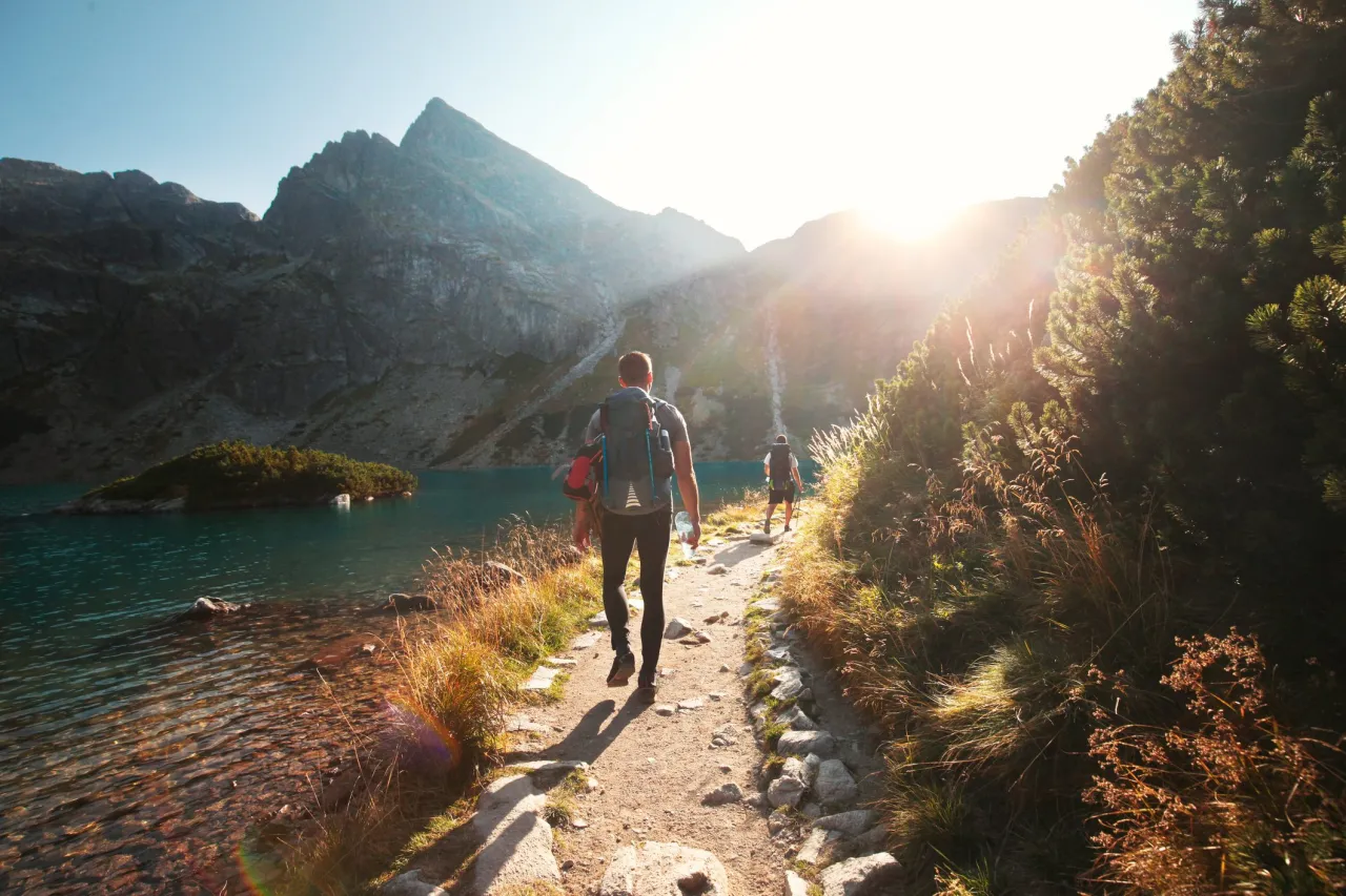 Turyści idą ścieżką nad jeziorem w g&oacute;rach. Słońce przebija się przez chmury, tworząc magiczny widok. To jedno z polska najpiękniejsze miejsca.