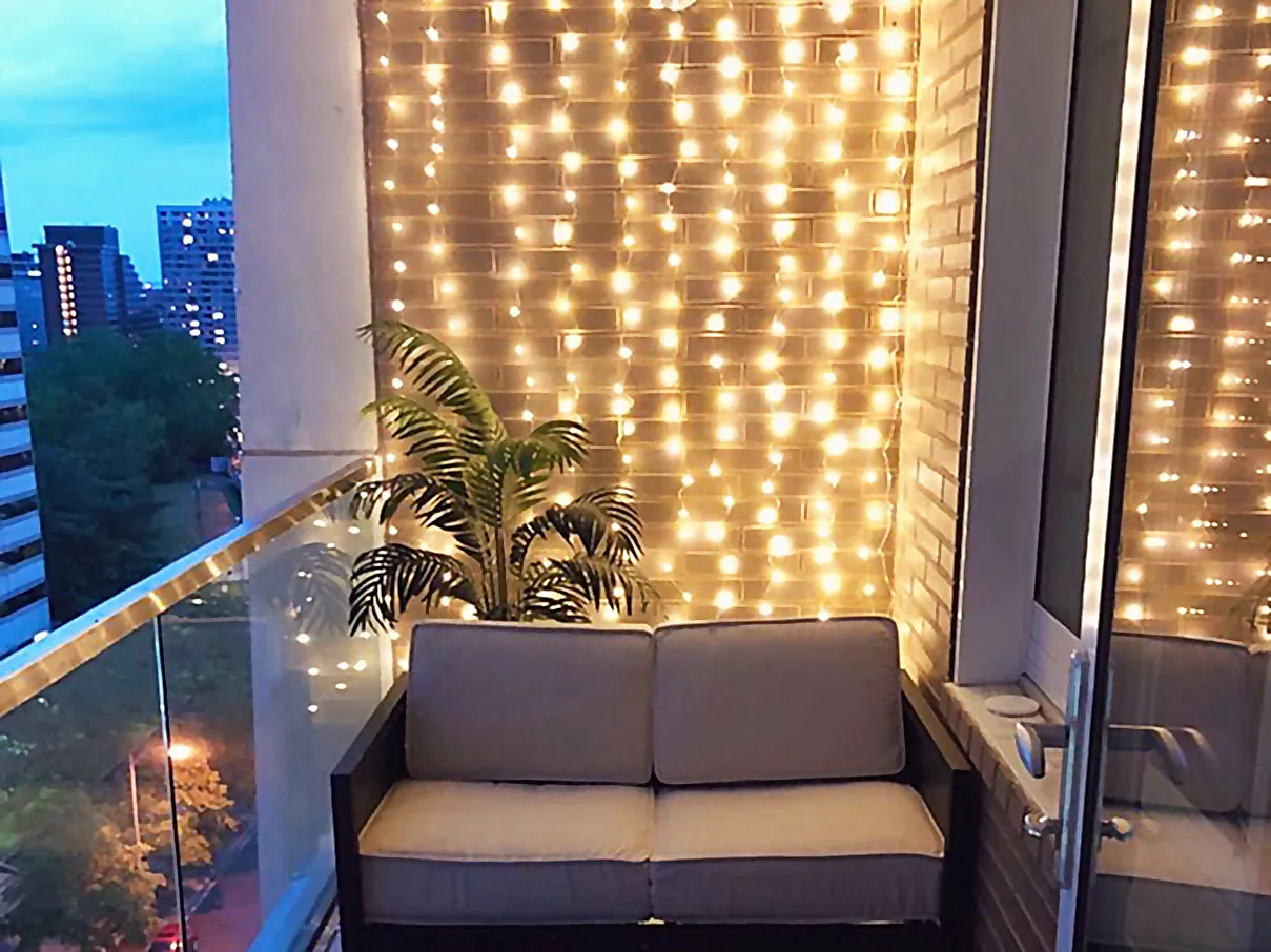 Uroczy balkon z sofą i palmą, ozdobiony ciepłym światłem lampek – idealny przykład, jak zrobić oświetlenie na balkonie, tworząc przytulną atmosferę.