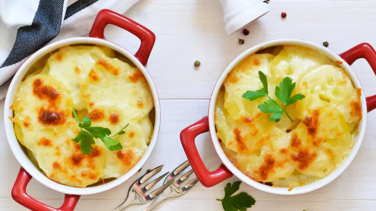 Dwie zapiekanki ziemniaczane w czerwonych naczyniach, z ciągnącym się serem i natką pietruszki. Idealne, gdy zastanawiasz się, z czego zrobić sos do zapiekanki ziemniaczanej.