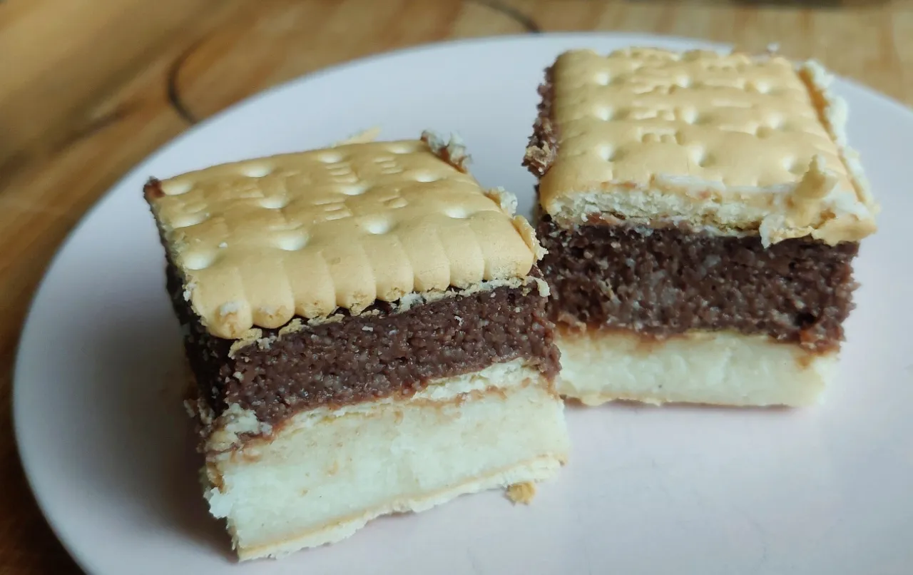 Dietetyczne ciasto z kaszy manny: warstwy biszkoptu, kremu czekoladowego i jasnego kremu, przykryte herbatnikami.
