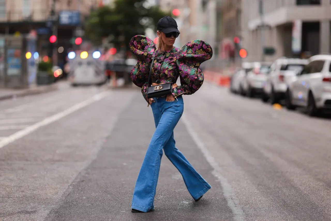 Kobieta w dzwonach i kwiecistej bluzce z bufiastymi rękawami idzie ulicą. Co do dzwonów, to jej stylizacja jest odważna i przyciąga wzrok.