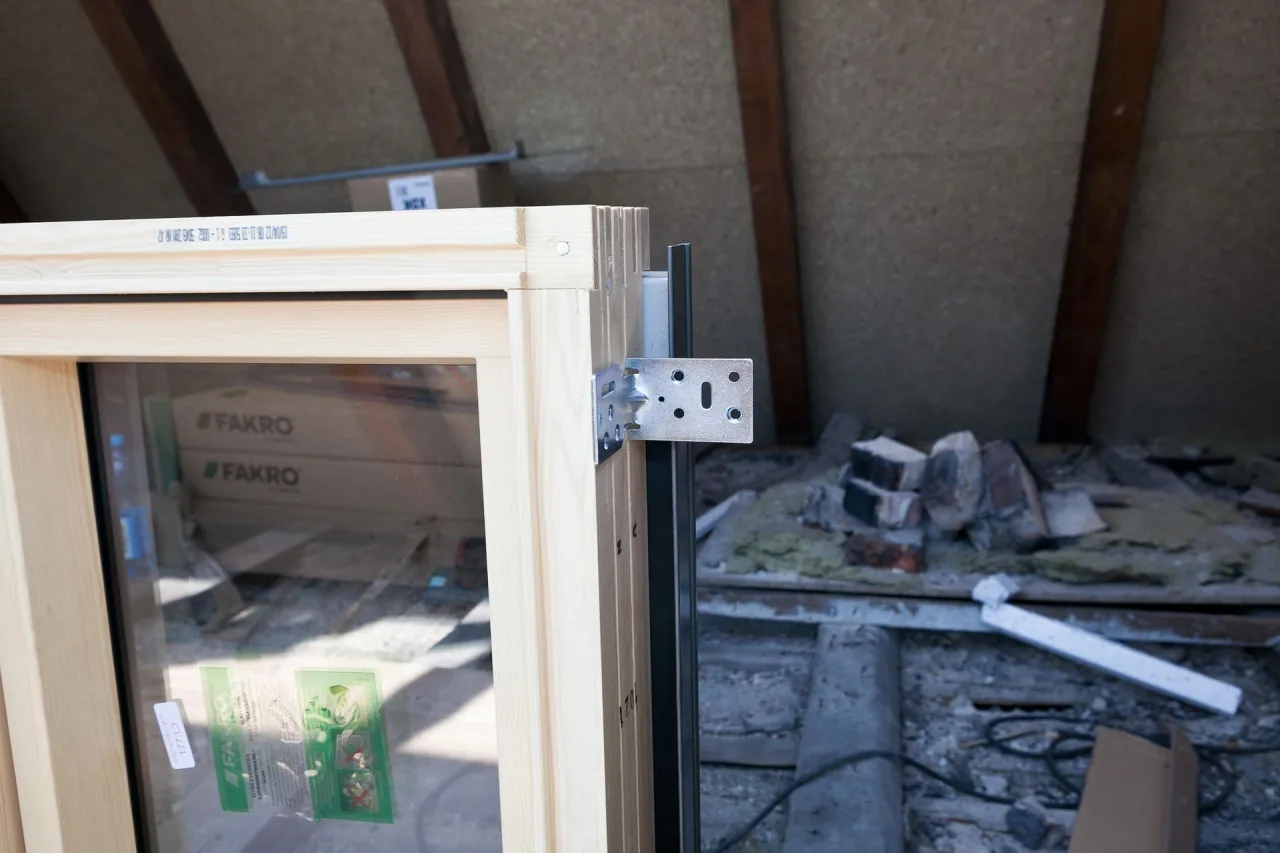 Drewniane okno FAKRO z metalowym kątownikiem, gotowe do ciepły montaż po zamontowaniu okien. W tle widać materiały budowlane.