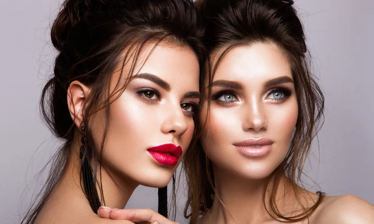 Dwie kobiety z perfekcyjnym makijażem, pokazujące, jak rozświetlić twarz. Jedna z nich ma wyraziste czerwone usta, druga delikatny błyszczący połysk.
