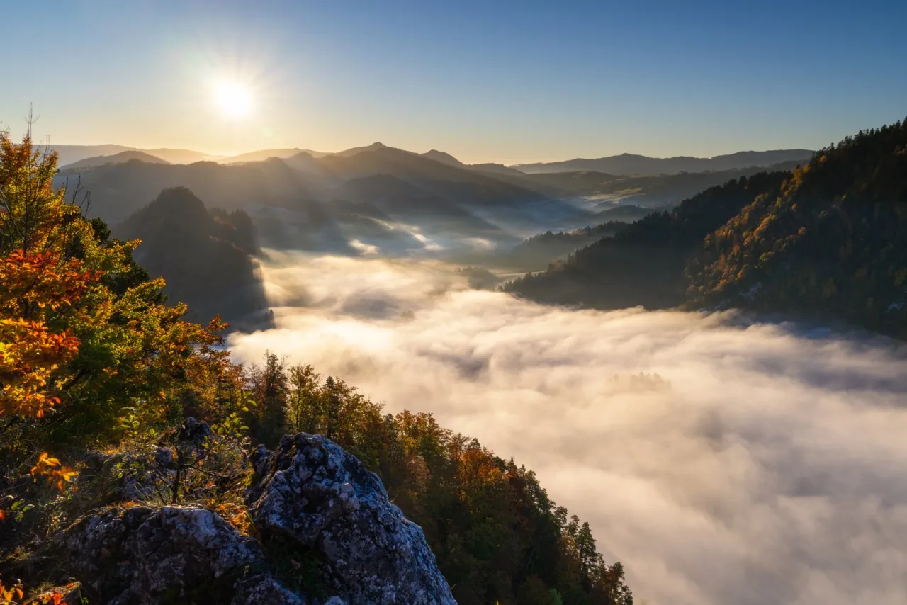 Mglisty poranek na szlakach w Tatrach, gdzie słońce przebija się przez chmury, oświetlając jesienne drzewa i skaliste zbocza.
