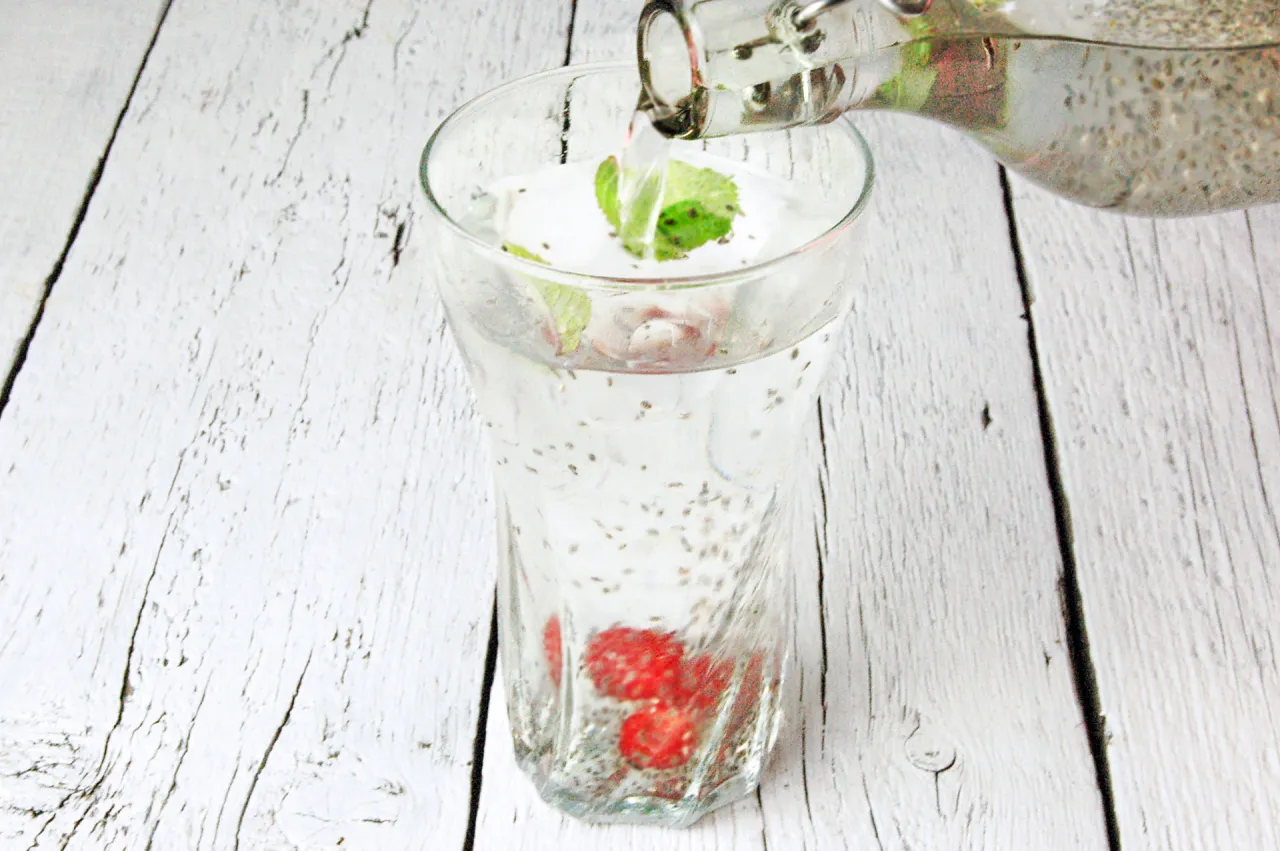 Jak pić nasiona chia? Woda z nasionami chia, malinami i miętą w szklance, nalewana z butelki.