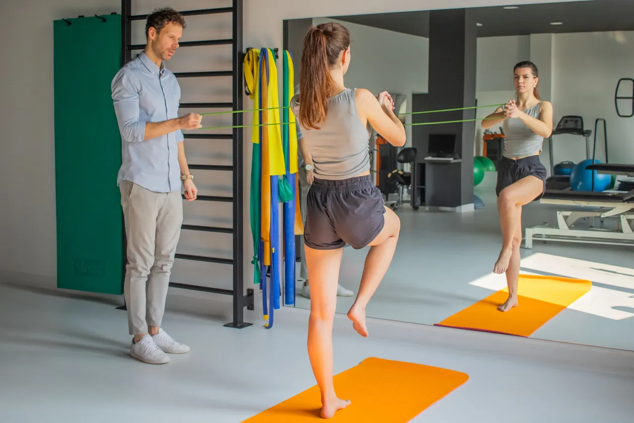 Fizjoterapeuta demonstruje ćwiczenia z gumą, jak wzmocnić kolana. Kobieta wykonuje ćwiczenie na macie.