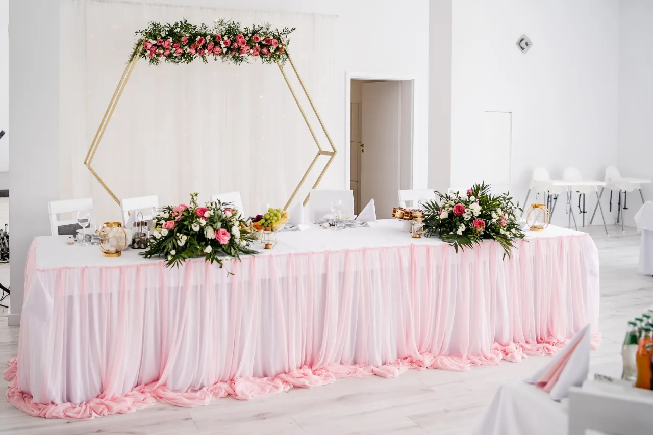 Elegancka dekoracja sali weselnej: złota konstrukcja z kwiatami, różowy tiulowy obrus i bukiety róż. Inspiracje na przyjęcie.